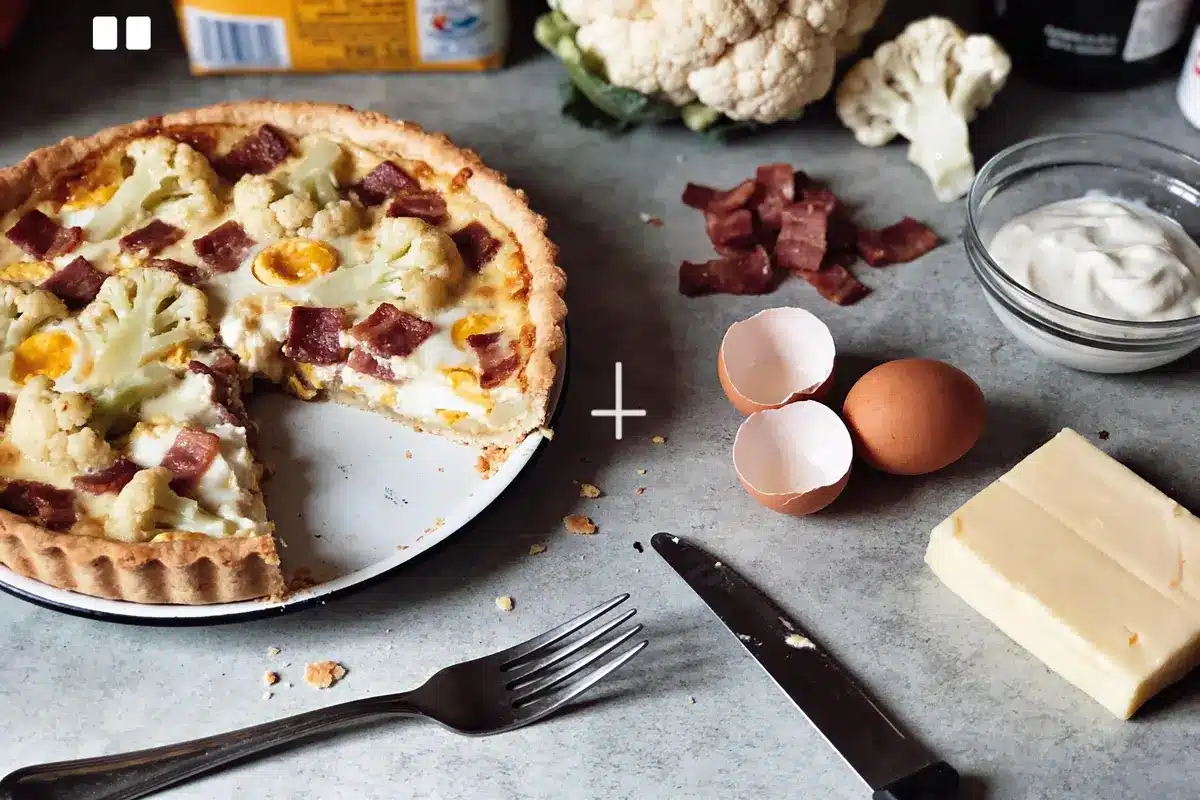 Quiche avec chou-fleur et salami, ingrédients environnants : œufs, fromage, crème, couteau et fourchette.