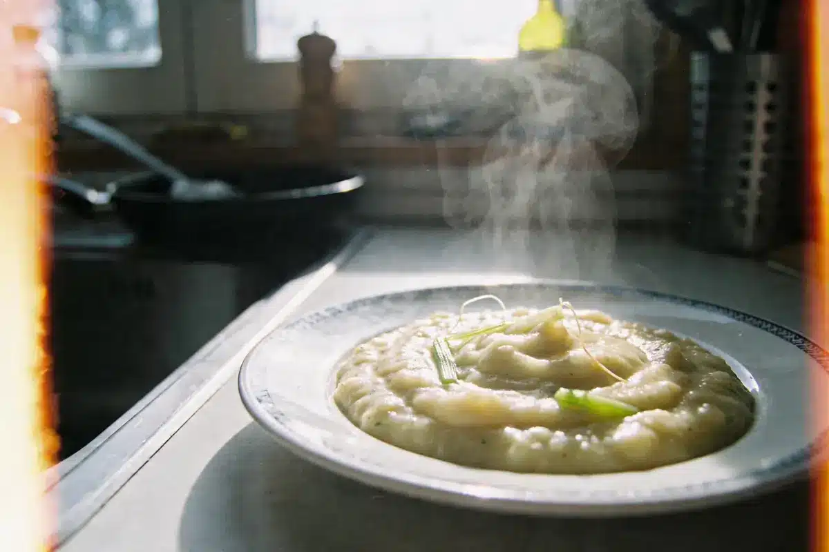 Assiette de purée fumante avec herbes, cuisine en arrière-plan, lumière naturelle douce.