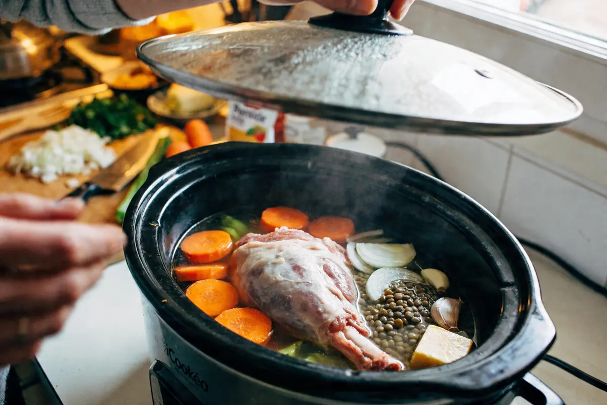 Slow cooker avec gigot d'agneau, carottes, lentilles, oignons et légumes. Couvercle tenu par une main.