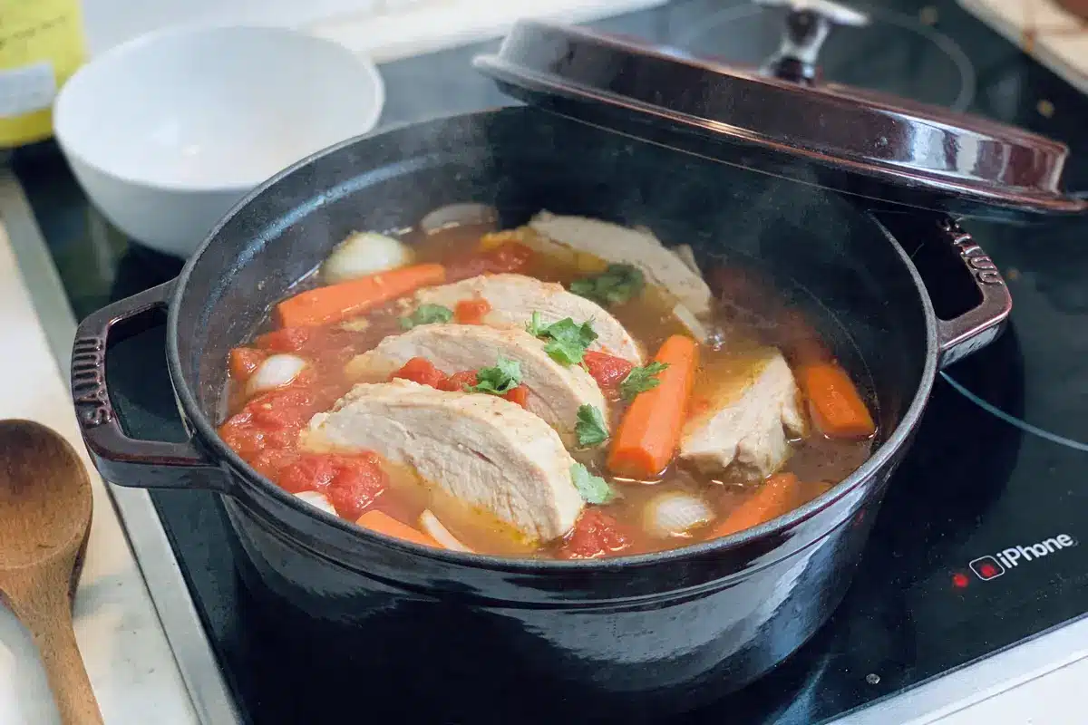 Cocotte en fonte sur plaque chauffante avec poulet, carottes, oignons, tomates et herbes, près d'une cuillère en bois.
