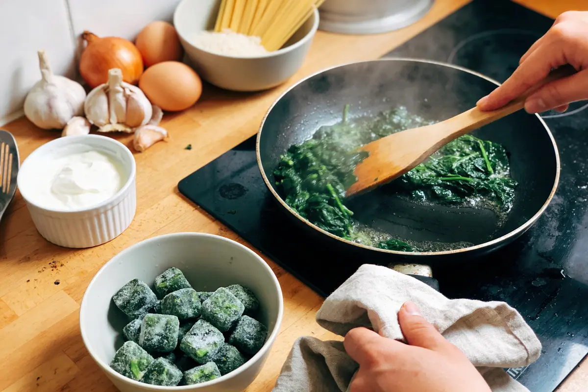 Personne cuisinant des épinards frais à la poêle, avec ingrédients variés sur le plan de travail.