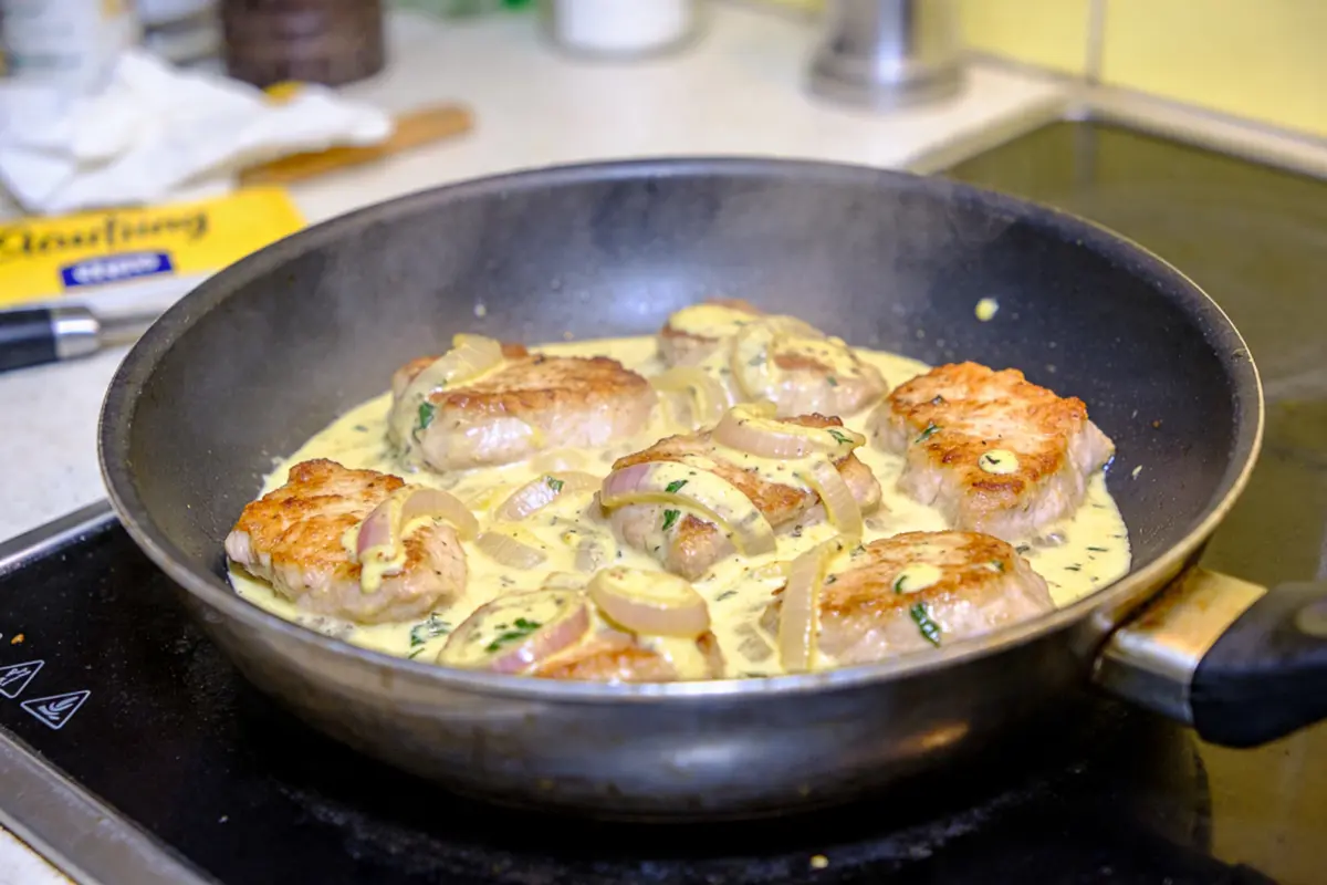 Morceaux de viande dans une poêle avec sauce crémeuse et rondelles d'oignon sur une cuisinière.