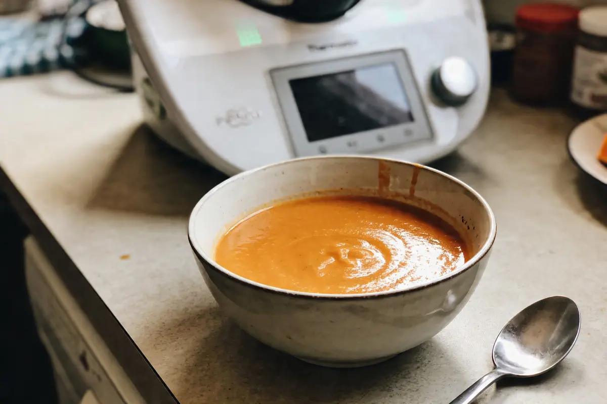 Bol de soupe orangée sur un plan de travail avec cuillère et robot culinaire en arrière-plan.