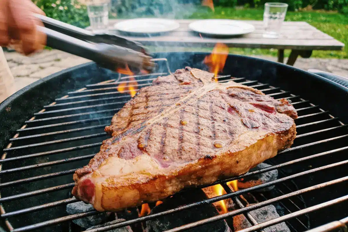 Viande grillée sur barbecue, avec flammes, pinces de cuisson, jardin en arrière-plan, table de pique-nique.