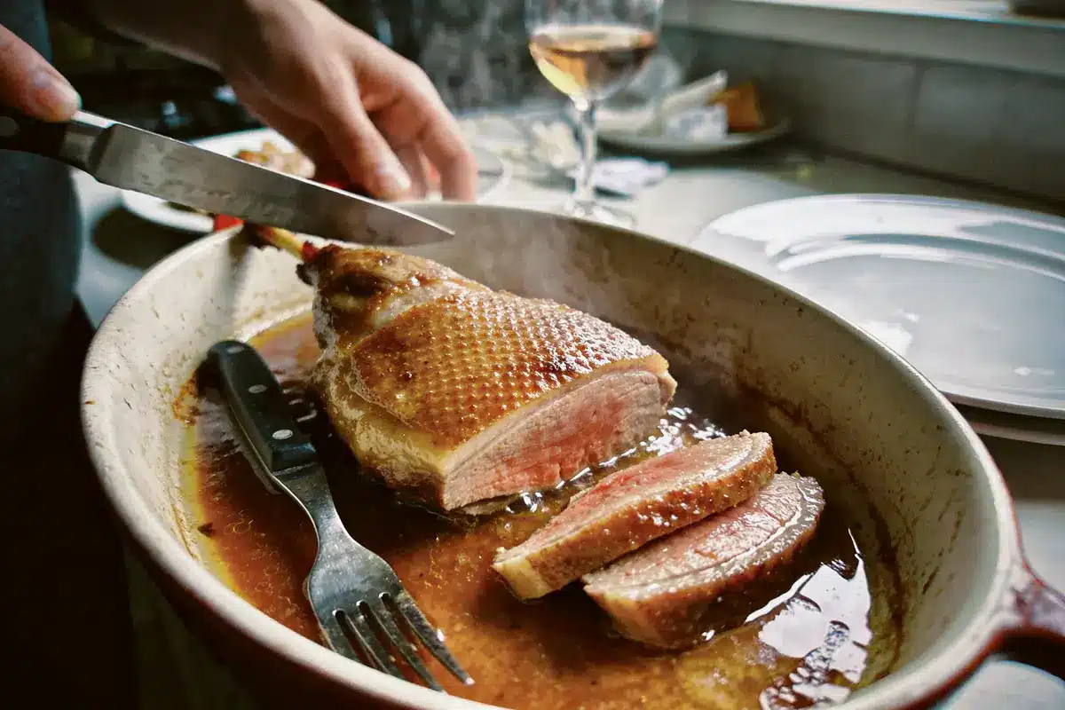 Personne tranchant du canard rôti dans un plat, verre de vin à côté.