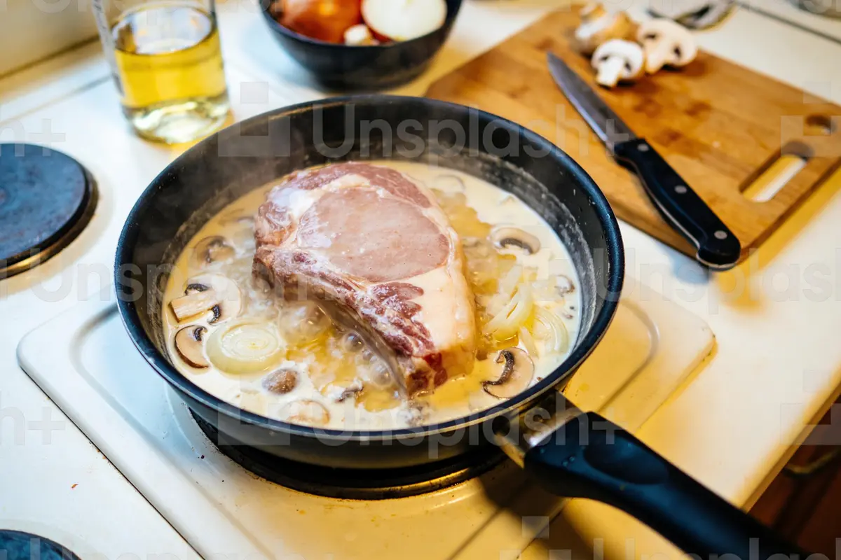 Côte de porc cuisant dans une poêle crémeuse avec champignons et oignons, planche de découpe à proximité.