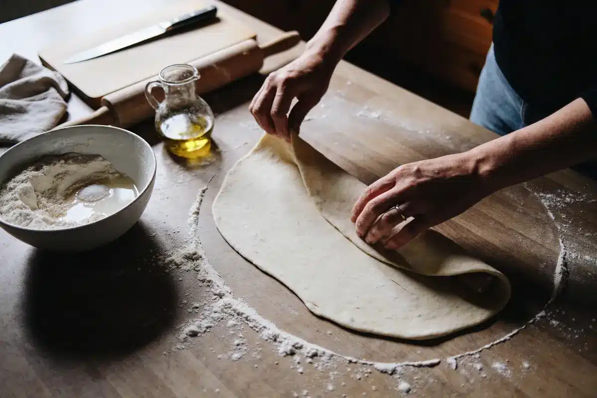 Personne préparant une pâte sur une table en bois, avec farine, huile d'olive, planche et couteau.