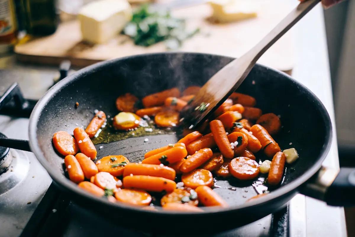Carottes sautées dans une poêle avec du beurre et des herbes, remuées avec une cuillère en bois.