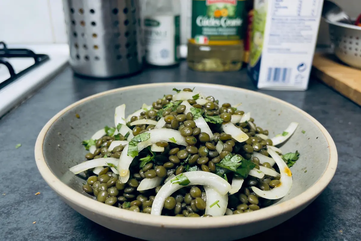 Bol de lentilles vertes avec oignons et persil, sur comptoir de cuisine avec divers ingrédients en arrière-plan.