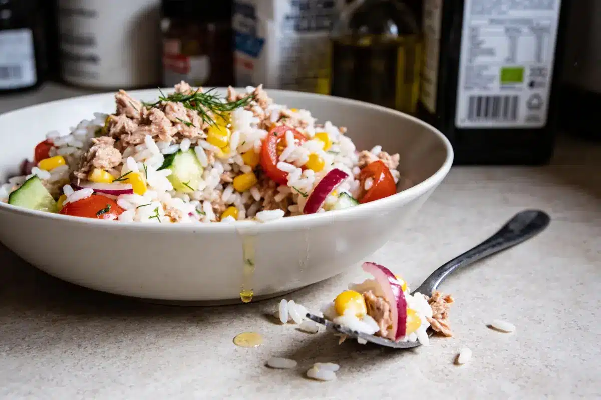 Salade de riz avec thon, tomates, concombre, maïs, oignons, dans un bol blanc, cuillère à côté.
