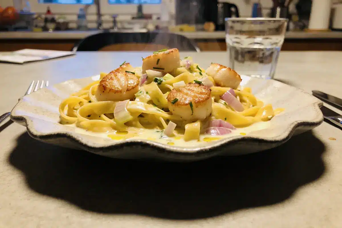 Assiette de tagliatelles crémeuses aux noix de Saint-Jacques et oignons rouges dans une cuisine.